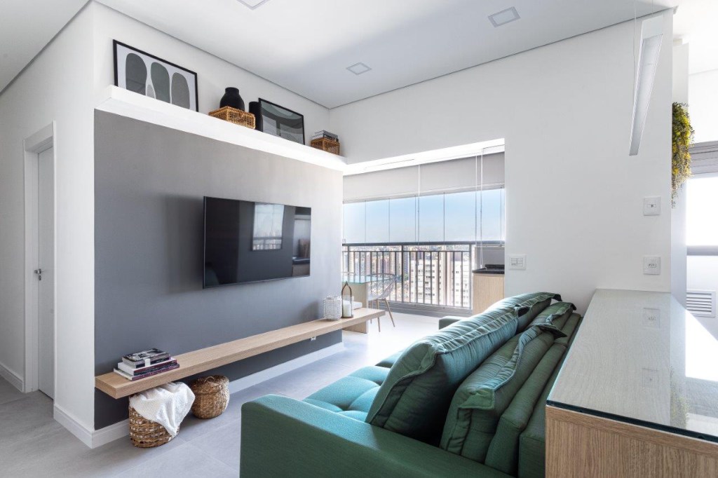 Sofá retrátil verde é destaque na sala de estar deste apartamento de 67 m². Projeto de Ju Miranda. Na foto, sala de estar, tv, parede cinza, sofá verde, prateleira branca.