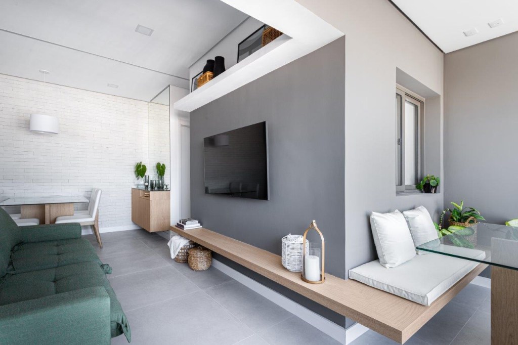 Sofá retrátil verde é destaque na sala de estar deste apartamento de 67 m². Projeto de Ju Miranda. Na foto, sala de estar, tv, parede cinza, sofá verde, prateleira branca. Banco fixo de madeira.