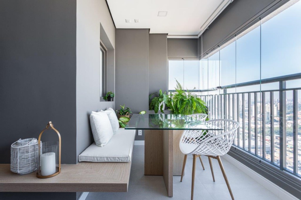 Sofá retrátil verde é destaque na sala de estar deste apartamento de 67 m². Projeto de Ju Miranda. Na foto, varanda fechada com vidro, banco fixo de madeira, futon, mesa com tampo de vidro, plantas.