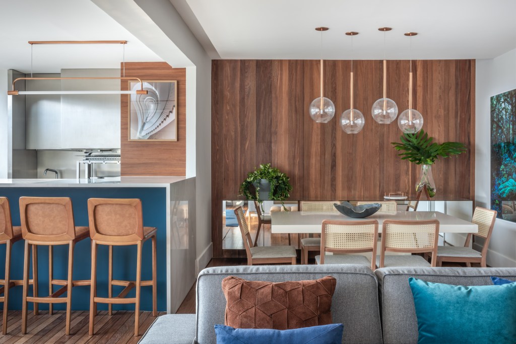 Apartamento ganha décor com tons de azul, cinza e madeira. Projeto de Beatriz Quinelato. Na foto, sala de jantar integrada com a varanda. Parede de madeira e sofá.