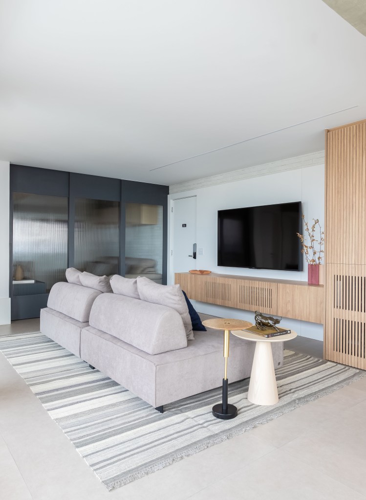 Apê de 120m² ganha cozinha integrada azul e sala de TV com porta de correr. Projeto de Carolina Gava. Na foto, sala de estar, tv, tapete listrado, sofá ilha cinza.