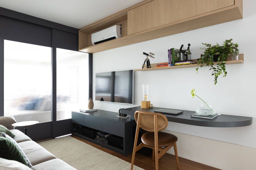 Apê de 120m² ganha cozinha integrada azul e sala de TV com porta de correr. Projeto de Carolina Gava. Na foto, home office, porta de vidro, sofá cinza, tv, ar condicionado.