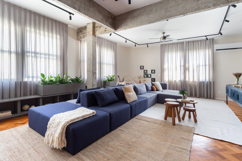 Apê de 192 m² mescla estilos industrial e brutalista com pitadas de cor. Projeto de Zimbro Arquitetura. Na foto, sala de estar com sofá ilha azul a pilares aparentes.