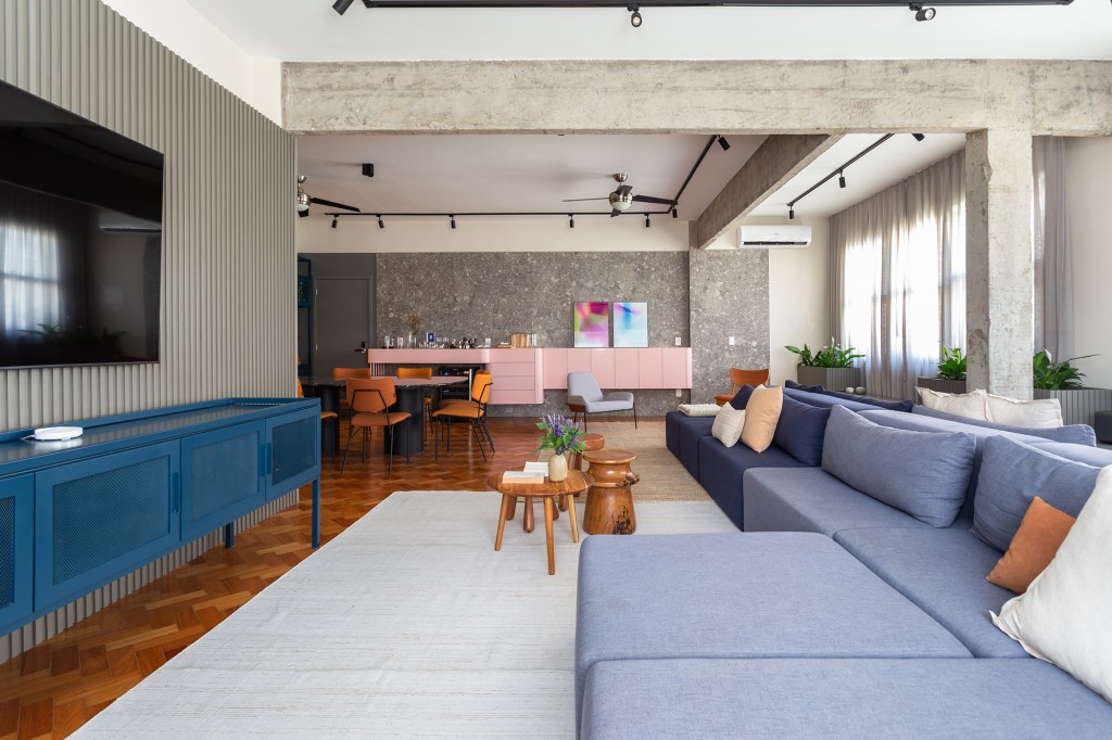Apê de 192 m² mescla estilos industrial e brutalista com pitadas de cor. Projeto de Zimbro Arquitetura. Na foto, sala de estar com tv, sofá azul e parede de concreto.