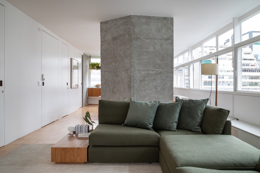 Apê dos anos 1960 é modernizado com dog shower e pilar no meio da sala. Projeto de Casa33 Arquitetura, Na foto, sala de estar com sofá verde, pilar na sala e mesas.