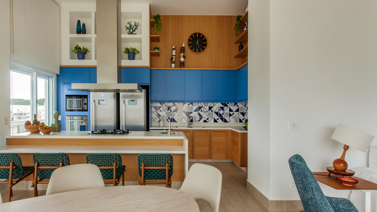 Apê tem décor inspirado na praia com toques azuis e porcelanato areia. Projeto de Korman Arquitetos. Na foto, cozinha integrada, mesa de jantar oval, backsplash de azulejos.