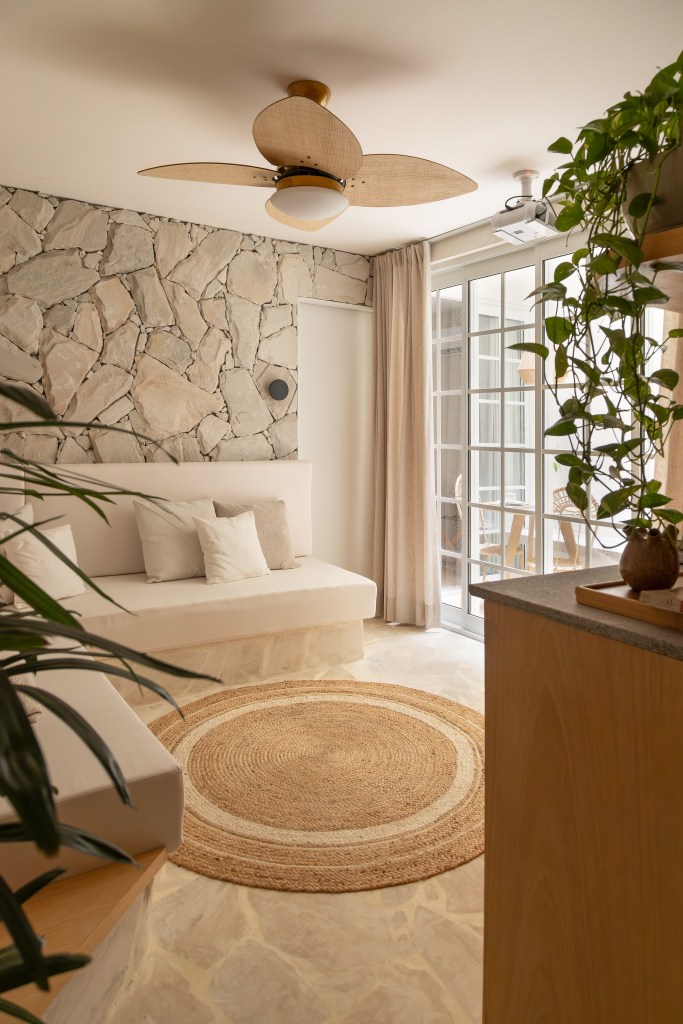 Apê térreo de 45 m² ganha cara de casa de praia com área externa integrada. Projeto de Casa Tauari. Na foto, sala de estar, tapete de fibra, parede de pedra, sofá branco, plantas.
