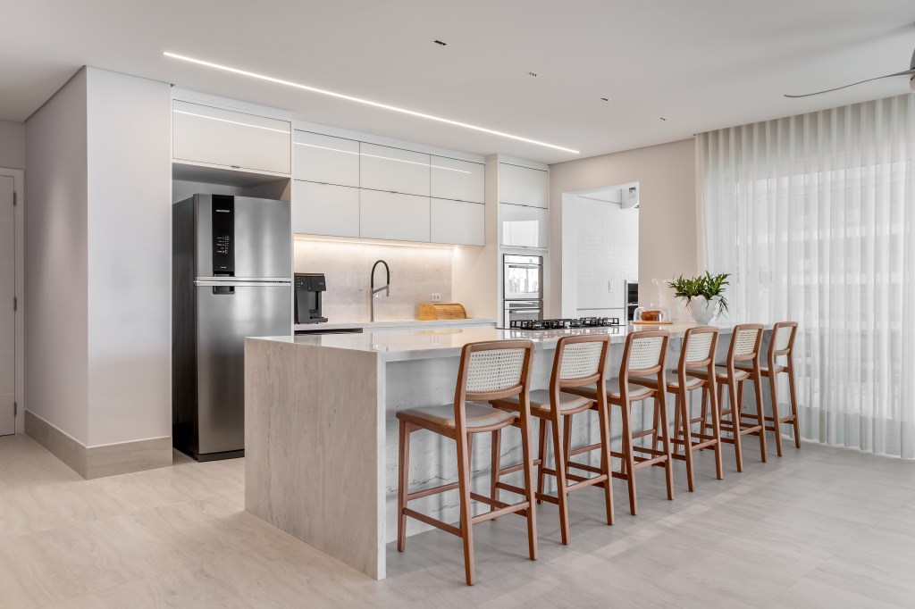 À beira-mar, apartamento de 160 m² traz uma estética clean e atemporal. Projeto de Mezzure Arquitetura. Na foto, cozinha americana como bancada. bancos e cortinas.