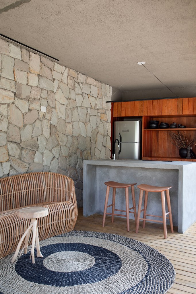 Chalé de praia de 60 m² tem decoração rústica e banheiro com claraboia. Projeto de Roby Macedo Arquitetura. Na foto, sala, cozinha integrada, bancada de pedra, banco de vime.