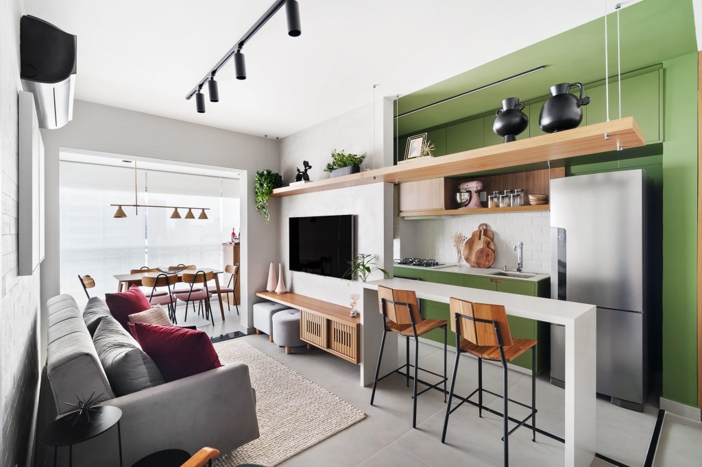 Com circulação otimizada, apê de 49 m² tem tons de verde e madeira. Projeto de Natalia Salla. Na foto, sala de estar integrada, sofá cinza, bancada branca, banquetas, cozinha. trilho de spots.