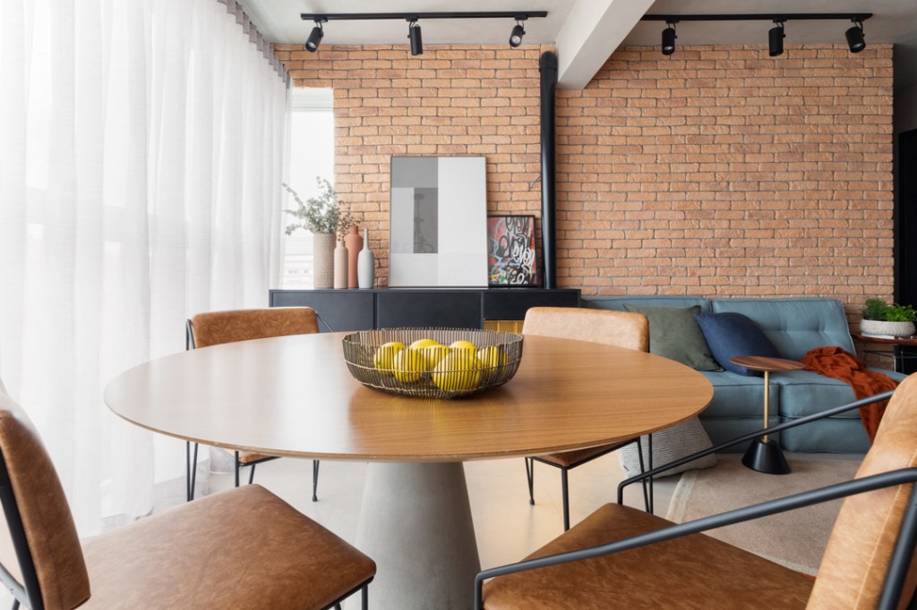 Concreto, tijolo aparente e integração dão ar industrial a apê de 72 m². Projeto de Studio Canto Arquitetura. Na foto, sala de estar e jantar com mesa redonda e sofá.