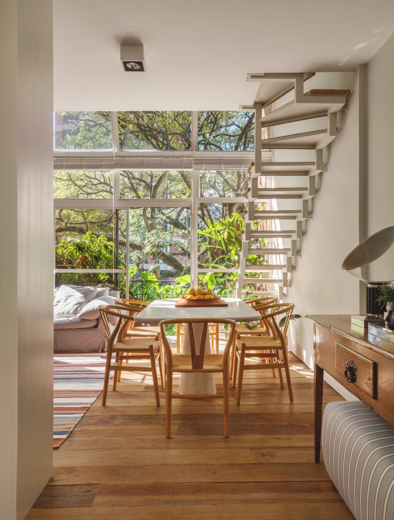 Mezanino é transformado em quarto nesta cobertura de 95 m². Projeto de DCC Arquitetura. Na foto, sala de estar, mesa de mármore, cadeiras de madeira, escada, grande janela com vista para árvores.