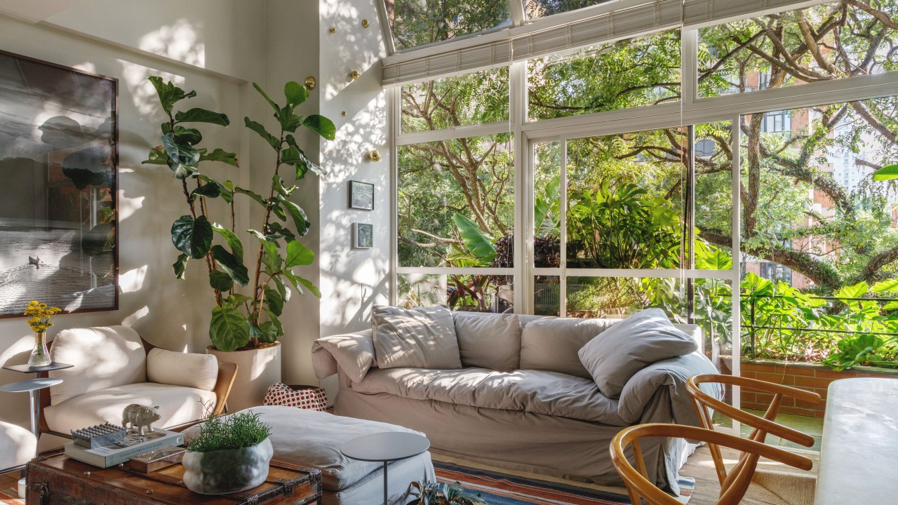 Mezanino é transformado em quarto nesta cobertura de 95 m². Projeto de DCC Arquitetura. Na foto, sala de estar, sofá cinza, grande janela com vista para árvores, tapete listrado, planta ficus.