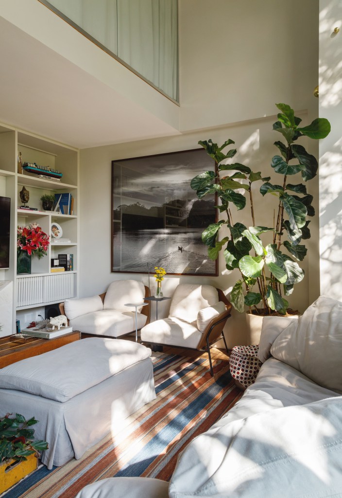 Mezanino é transformado em quarto nesta cobertura de 95 m². Projeto de DCC Arquitetura. Na foto, sala de estar, pufe, poltronas brancas, quadro, tapete listrado, planta ficus.
