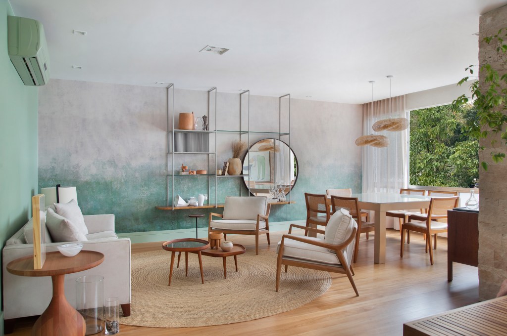 Papel de parede com efeito degradê envelhecido é destaque em área social. Projeto de Manoela Fleck. Na foto, sala de estar, mesa, poltronas brancas, espelho redondo, estante metálica vazada.