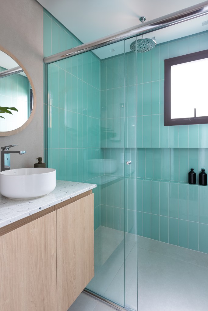 Reforma amplia living de apê de 100 m² com marcenaria planejada e plantas. Projeto de Estúdio Maré. Na foto, banheiro pequeno, espelho redondo, box com azulejos azuis.