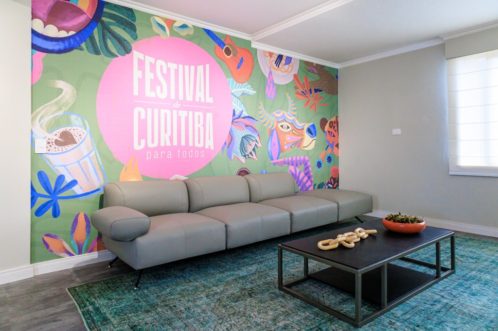 Sala é inspirada na obra de Zé Celso e na exuberância da Amazônia. Projeto de Jordana Fraga. Na foto, sala, papel de parede florido, sofá cinza, tapete.
