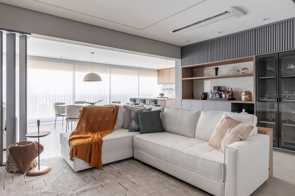 Varanda de apê tem dois espaços: um gourmet e outro de estar. Projeto de Blaia e Moura. Na foto, sala de estar integrada com varanda, sofá branco, bar.