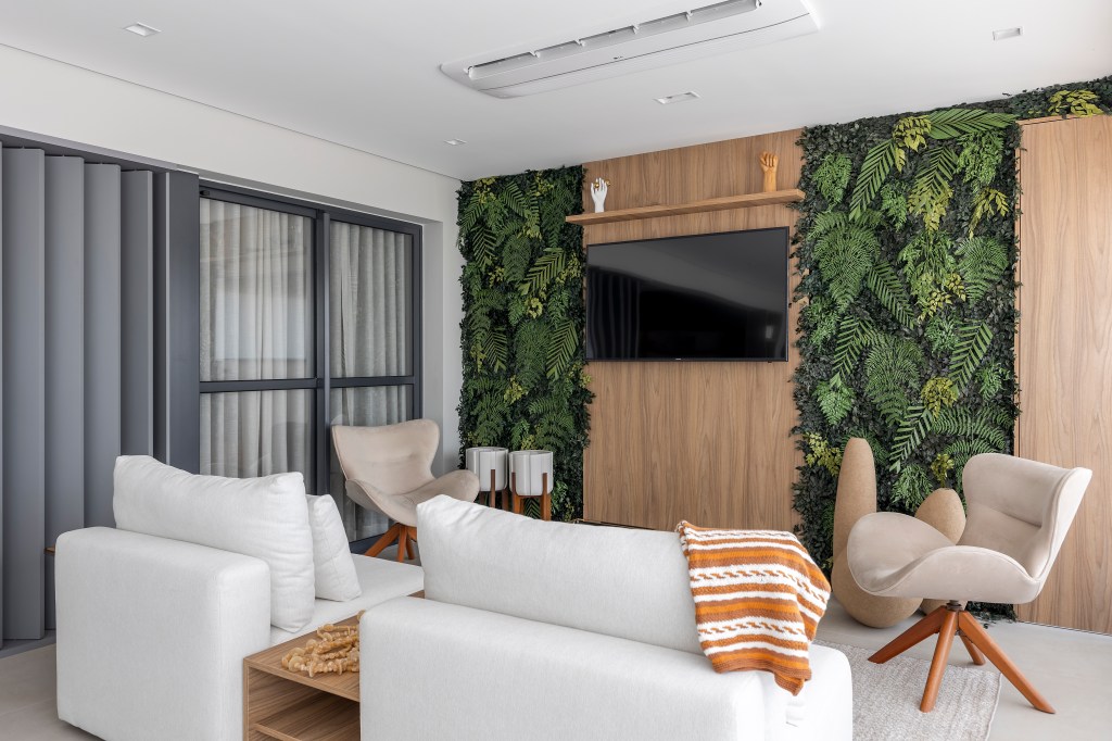 Varanda de apê tem dois espaços: um gourmet e outro de estar. Projeto de Blaia e Moura. Na foto, varanda com painel de tv, jardim vertical e sofá branco.