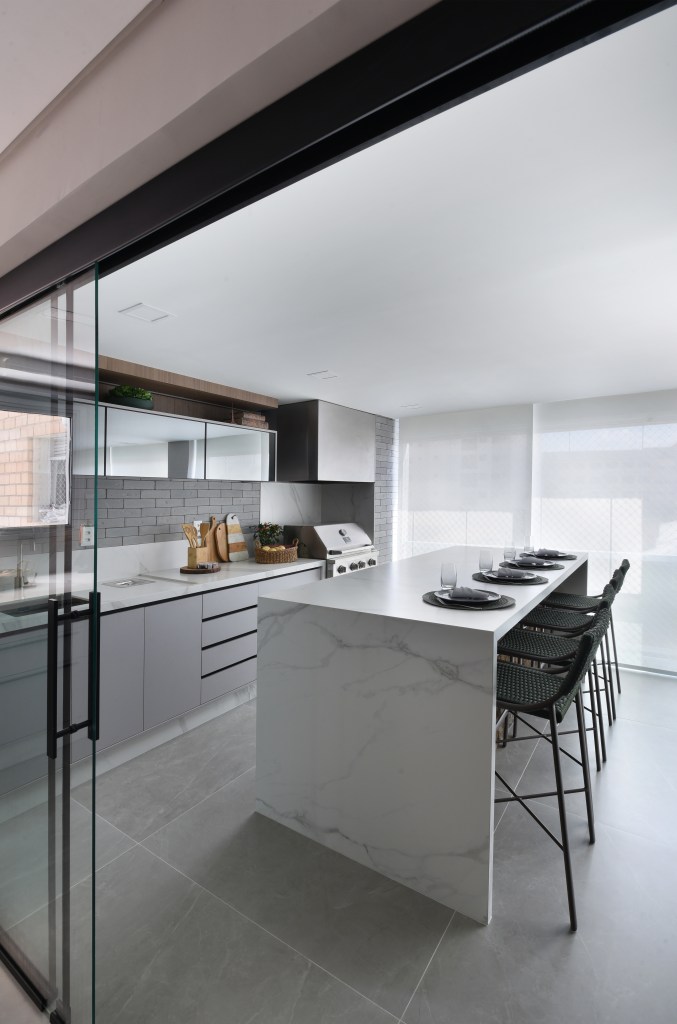 Varanda de estar gourmet é destaque em apartamento de 204 m². Projeto de Rosangela Pena. Na foto, cozinha com ilha e bancada marmorizada, banquetas, marcenaria cinza.