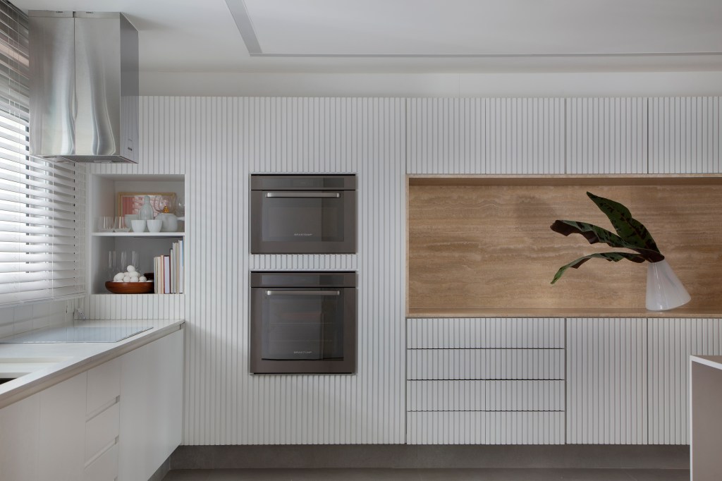 Venezianas em madeira trazem privacidade aos espaços de apê minimalista. Projeto de PKB Arquitetura. Na foto, cozinha com marcenaria ripada branca.