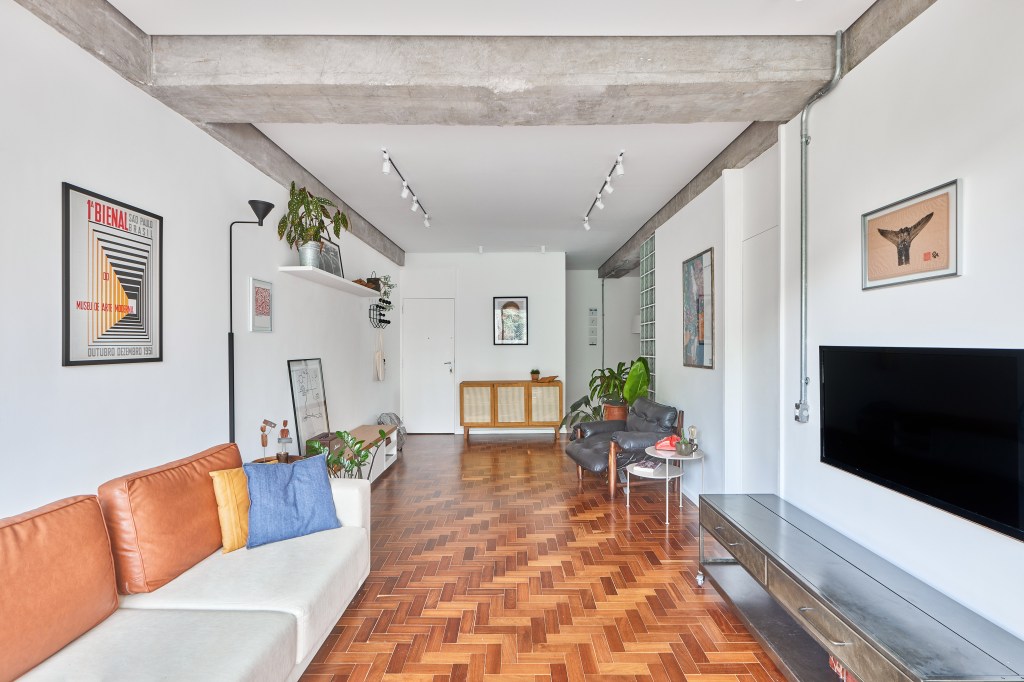 Vigas de concreto aparente emolduram área social de apê de 98 m². Projeto de Hugo Rapizo. Na foto, sala de estar, piso de taco, sofá branco, almofadas coloridas.