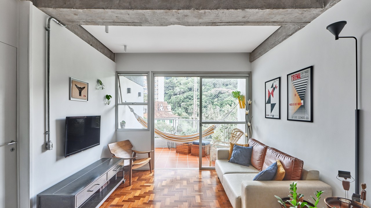 Vigas de concreto aparente emolduram área social de apê de 98 m². Projeto de Hugo Rapizo. Na foto, sala de estar, piso de taco, varanda com rede, sofá branco.