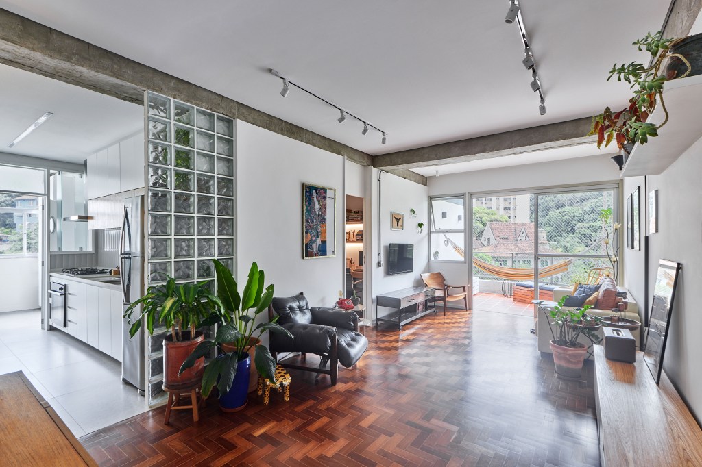 Vigas de concreto aparente emolduram área social de apê de 98 m². Projeto de Hugo Rapizo. Na foto, sala de estar, piso de taco, plantas, parede de tijolos de vidro.