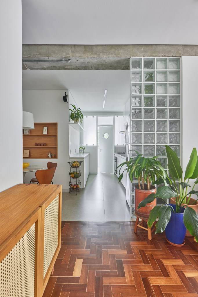 Vigas de concreto aparente emolduram área social de apê de 98 m². Projeto de Hugo Rapizo. Na foto, cozinha, plantas, parede com tijolos de vidro,