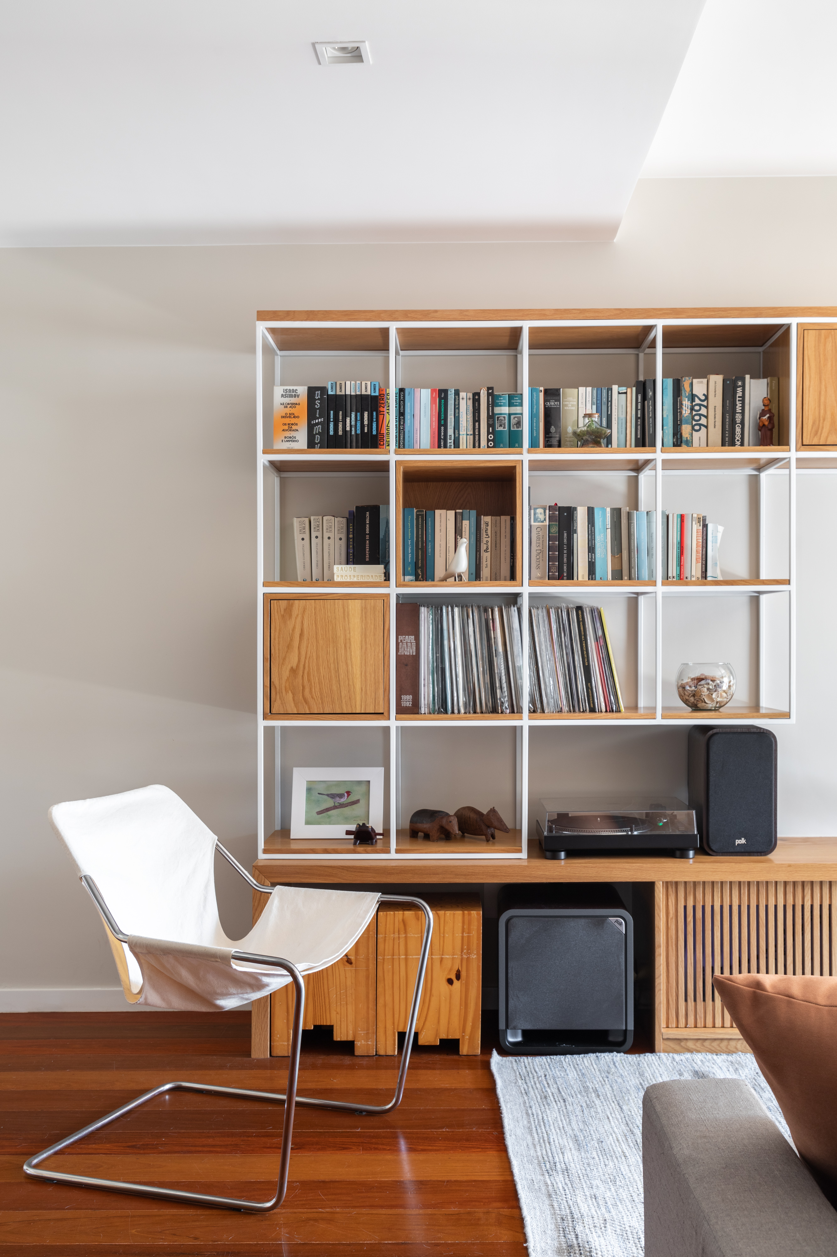 Apê ganha estante para livros e discos de vinil e paredes com ladrilhos. Projeto de Co+Lab Juntos Arquitetura. Na foto, sala, piso de madeira, poltrona branca.