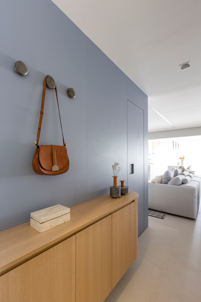 Cabideiro e sapateira: o conjunto que traz praticidade e beleza ao hall. Projeto de Mari Milani. Na foto, parede azul acinzentada, gancho, bolsa pequena.