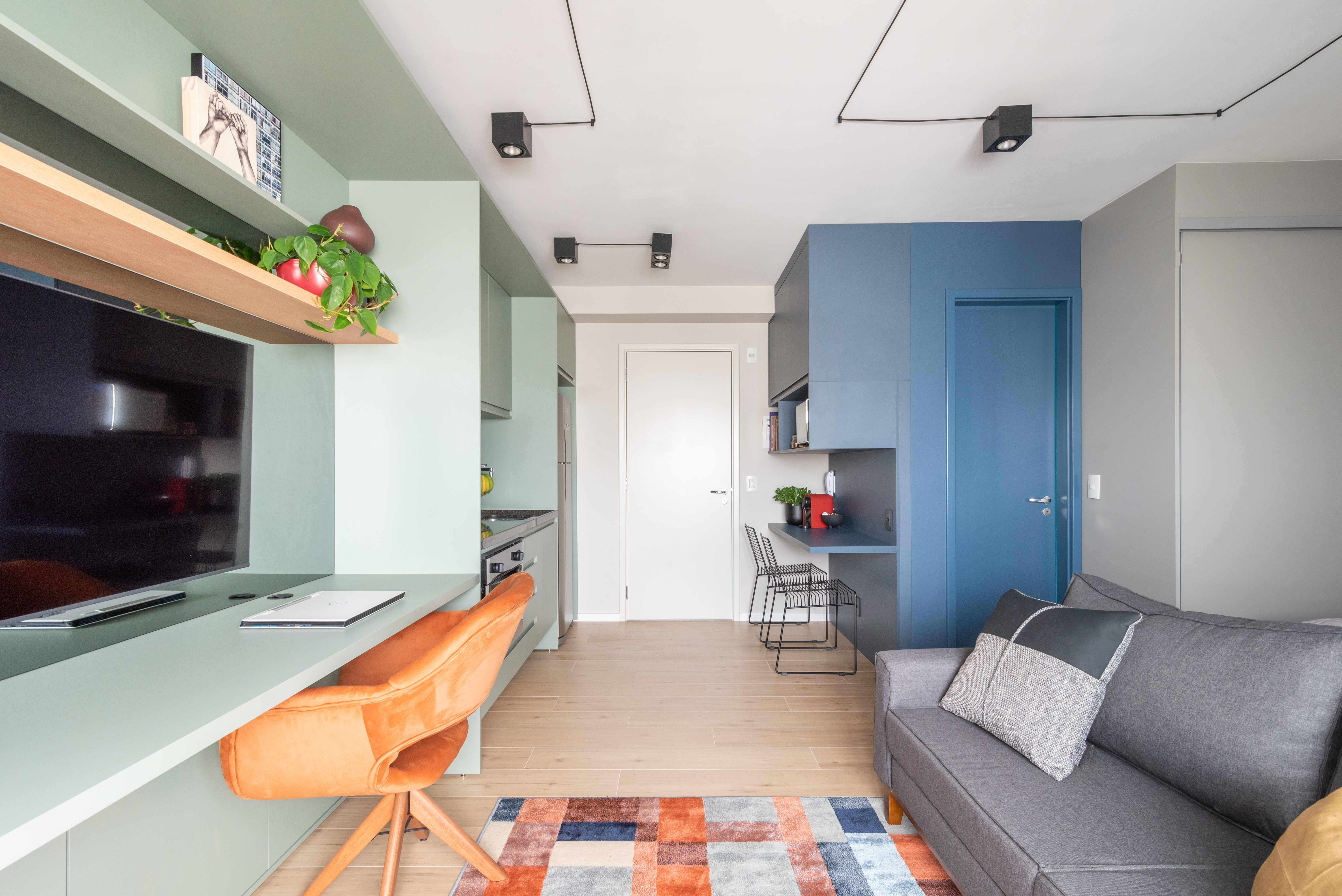 Caixa de marcenaria verde abriga cozinha, bancada e tv em estúdio de 34 m². Projeto de Abrazo Arquitetura. Na foto, sala, parede e marcenaria azul, poltrona, tapete quadriculado colorido.