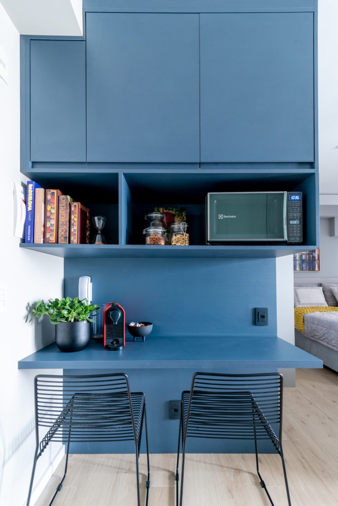 Caixa de marcenaria verde abriga cozinha, bancada e tv em estúdio de 34 m². Projeto de Abrazo Arquitetura. Na foto, marcenaria azul com bancada para refeições, duas cadeiras, cantinho do café.
