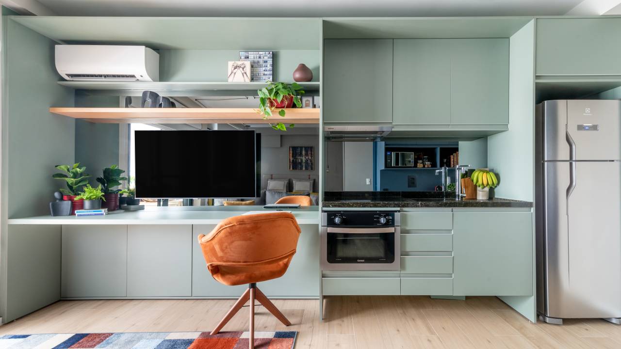 Caixa de marcenaria verde abriga cozinha, bancada e tv em estúdio de 34 m². Projeto de Abrazo Arquitetura. Na foto, bancada, poltrona, ar condicionado, tv, tapete quadriculado colorido.