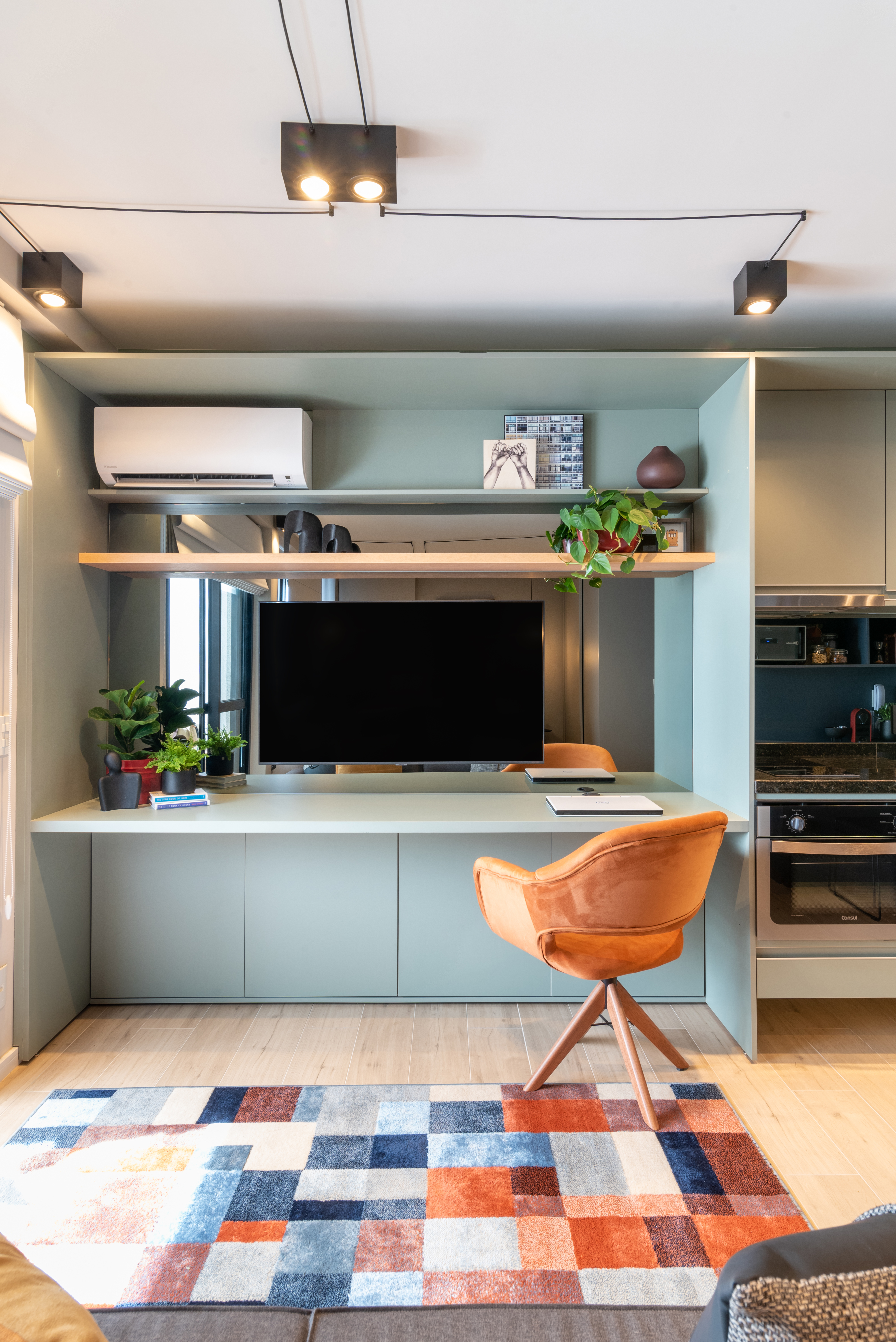 Caixa de marcenaria verde abriga cozinha, bancada e tv em estúdio de 34 m². Projeto de Abrazo Arquitetura. Na foto, bancada, poltrona, ar condicionado, tv, tapete quadriculado colorido.