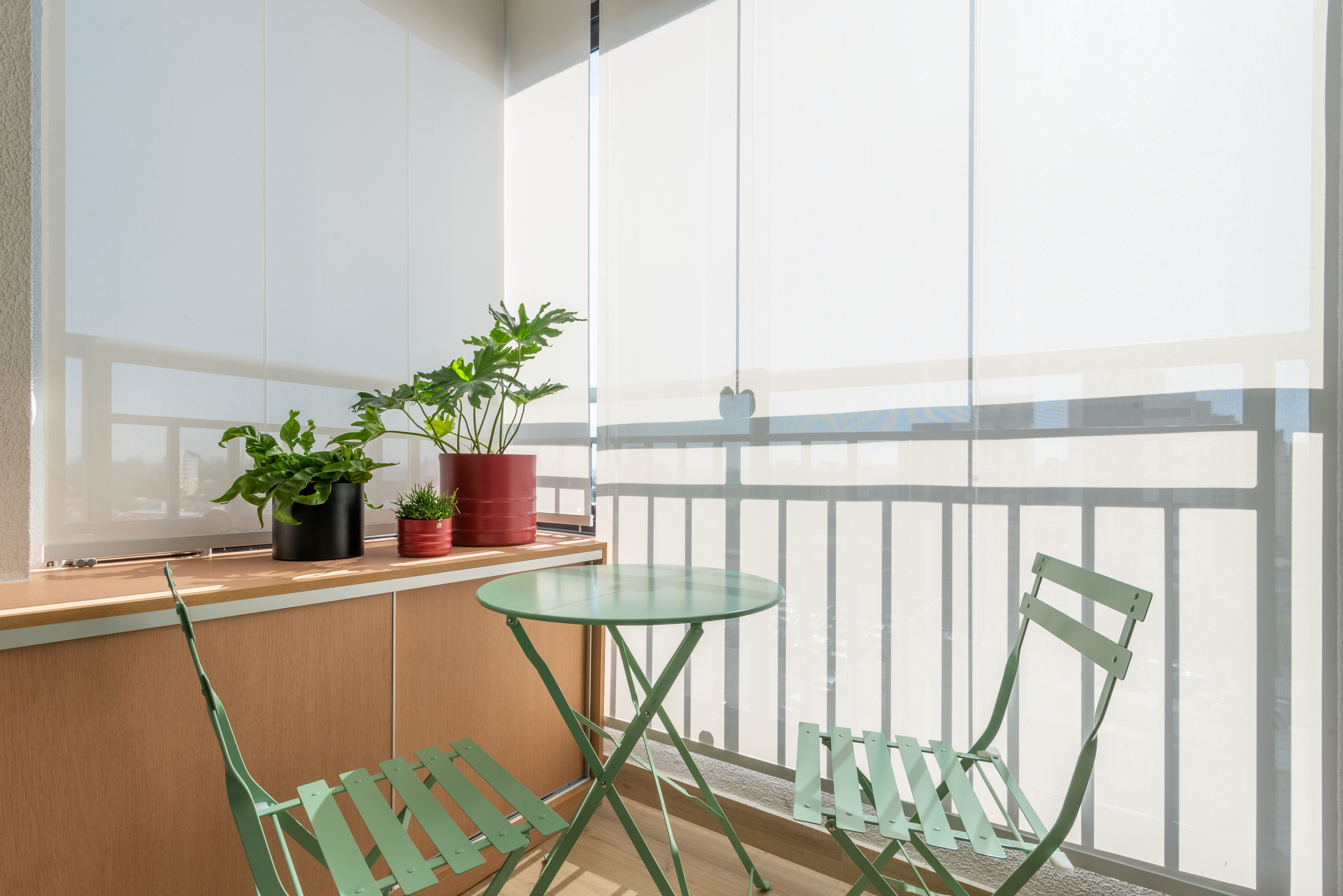 Caixa de marcenaria verde abriga cozinha, bancada e tv em estúdio de 34 m². Projeto de Abrazo Arquitetura. Na foto, varanda com mesa pequena e cadeiras verdes.