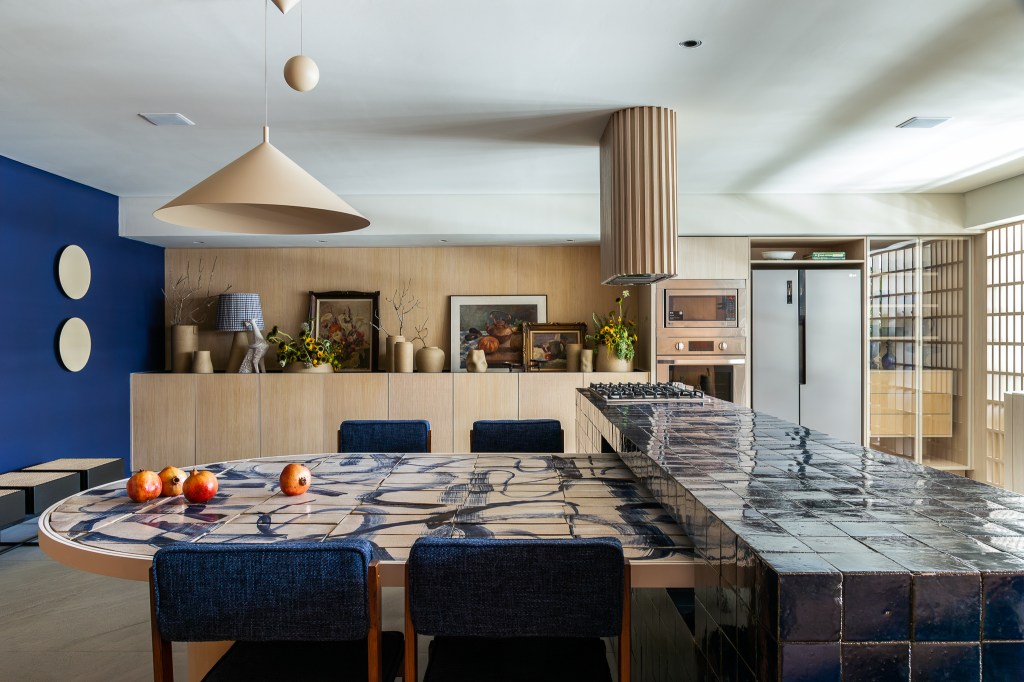Cerâmicas artesanais compõem o tampo da mesa desta cozinha repleta de azul. Projeto de Mandril Arquitetura. Na foto, cozinha com bancada azul e mesa estampada de cerâmica.