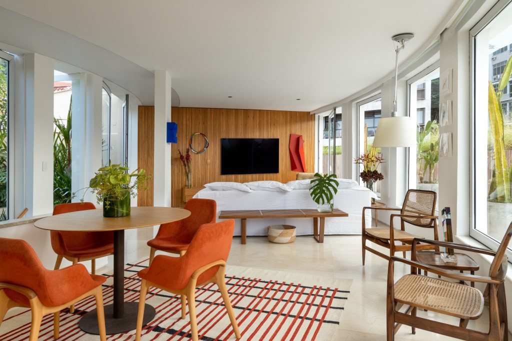 Cobertura com planta circular tem jardim interno, área gourmet e sauna. Projeto de Roberto Souto. Na foto, sala integrada, sofá branco, banco, mesa redonda, cadeiras laranja.