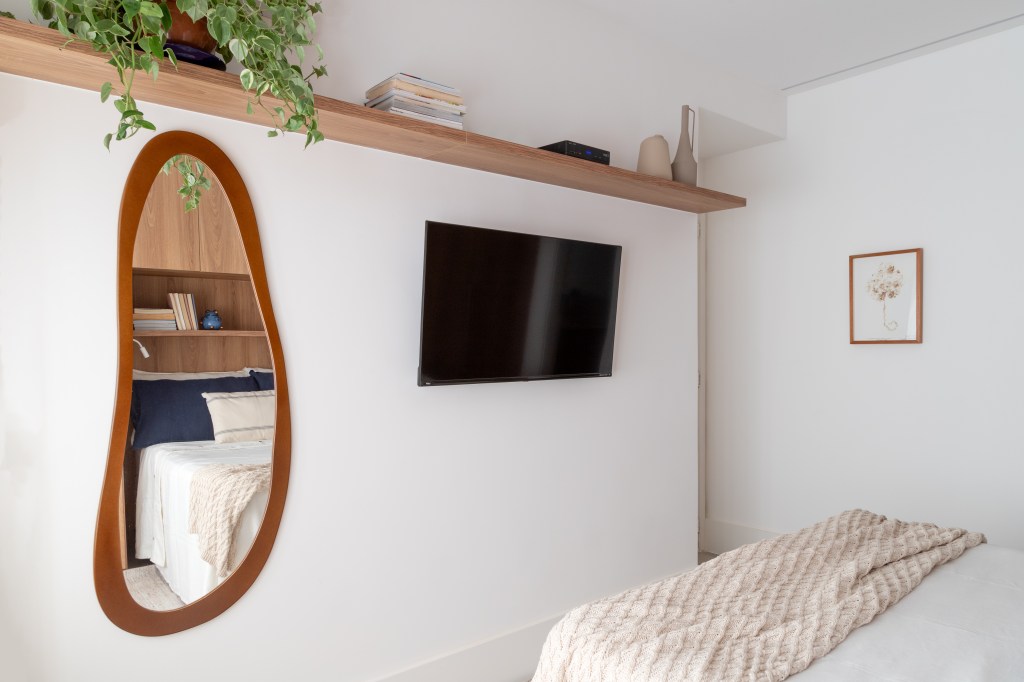 Espelho oval e armário em volta da cama são destaques em apê de 67 m². Projeto de Mariana Monnerat. Na foto, quarto, tv, prateleira.