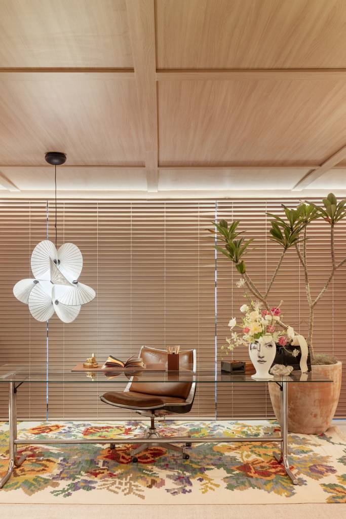 Home office de 45 m² tem estante de marcenaria e mármore e área de estar. Projeto de Rodolfo Consoli para a CASACOR São Paulo 2024. Na foto, mesa tampo de vidro, luminária, persiana de madeira.
