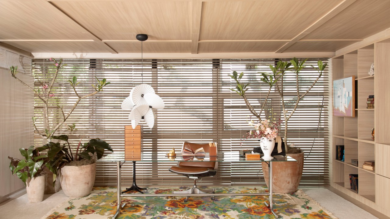 Home office de 45 m² tem estante de marcenaria e mármore e área de estar. Projeto de Rodolfo Consoli para a CASACOR São Paulo 2024. Na foto, mesa tampo de vidro, luminária, persiana de madeira.