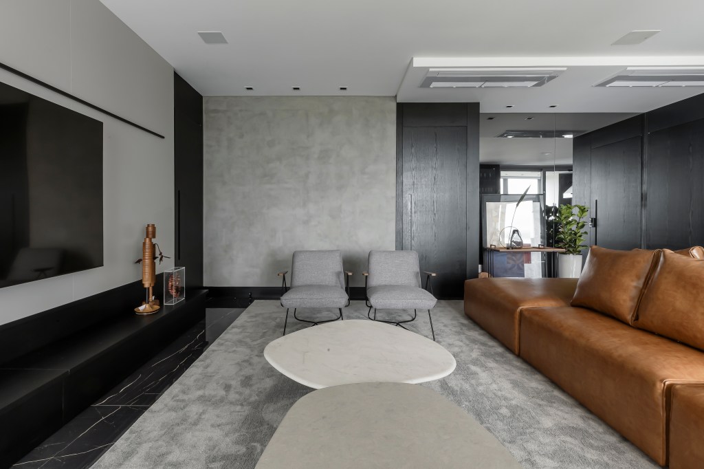 Móveis soltos e paleta sóbria deixam apê de 190 m² atemporal e elegante. Projeto de NK Arquitetura. Na foto, sala de estar cinza, sofá de couro, poltronas, tapete, mesas de centro em formato orgânico.