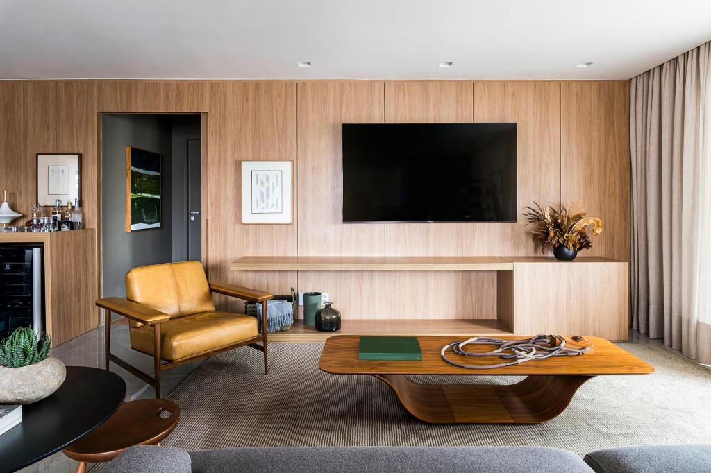 Em um mês, apê alugado de 156 m² ganha projeto contemporâneo e minimalista. Projeto Estúdio Amadas. Na foto, sala de TV com parede de madeira, mesa, tapete e poltrona.