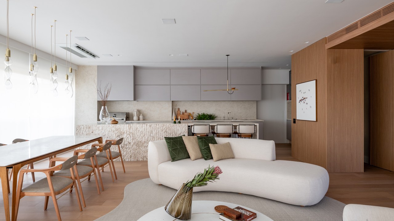 Vista 180º para o Ibirapuera e banheiro SPA marcam este apartamento. Projeto de Ana Carolina Queiroz. Na foto, sala integrada com a cozinha, sofá curvo, bancada de pedra e mesa de jantar.