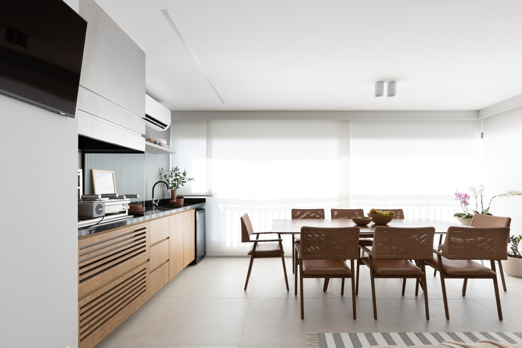 Aconchego e praticidade: apartamento de 126m² possui porcelanato acinzentado neutro e vinílico. Projeto do Estúdio Bena. Na foto, varanda gourmet com churrasqueira e mesa de jantar