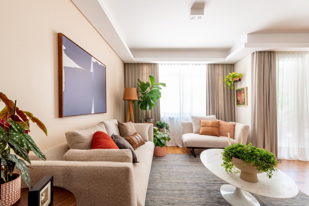 Apartamento térreo ganha brinquedoteca com papel de parede tropical. Projeto de Abrazo Interiores. Na foto, sala de estar, cortina, sofá bege, plantas, mesa de centro oval com tampo de mármore.