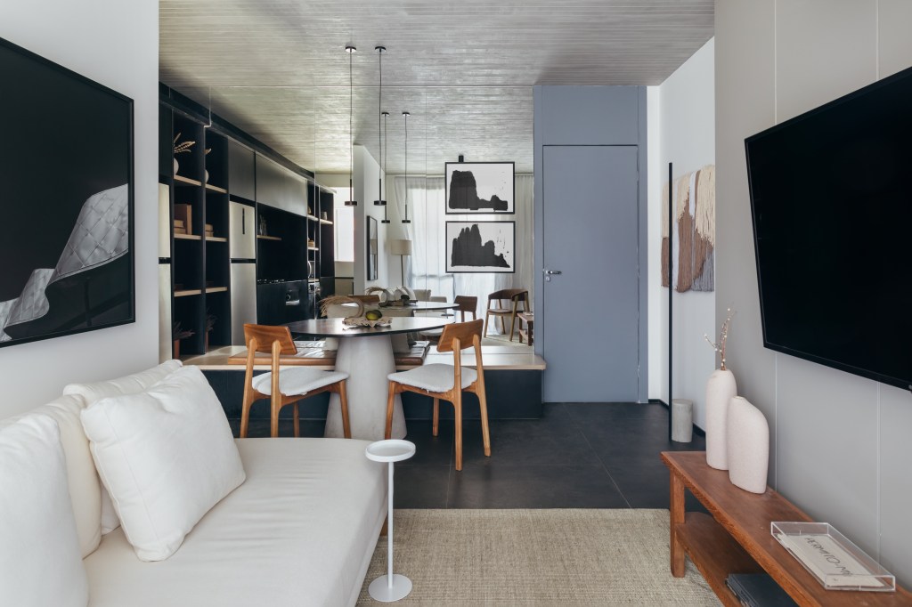 Apê de 73 m² tem marcenaria ebanizada e piso de porcelanato preto. Projeto de Studio Leandro Neves. Na foto, sala, parede preta, teto de concreto, sofá branco, mesa redonda, cozinha integrada.