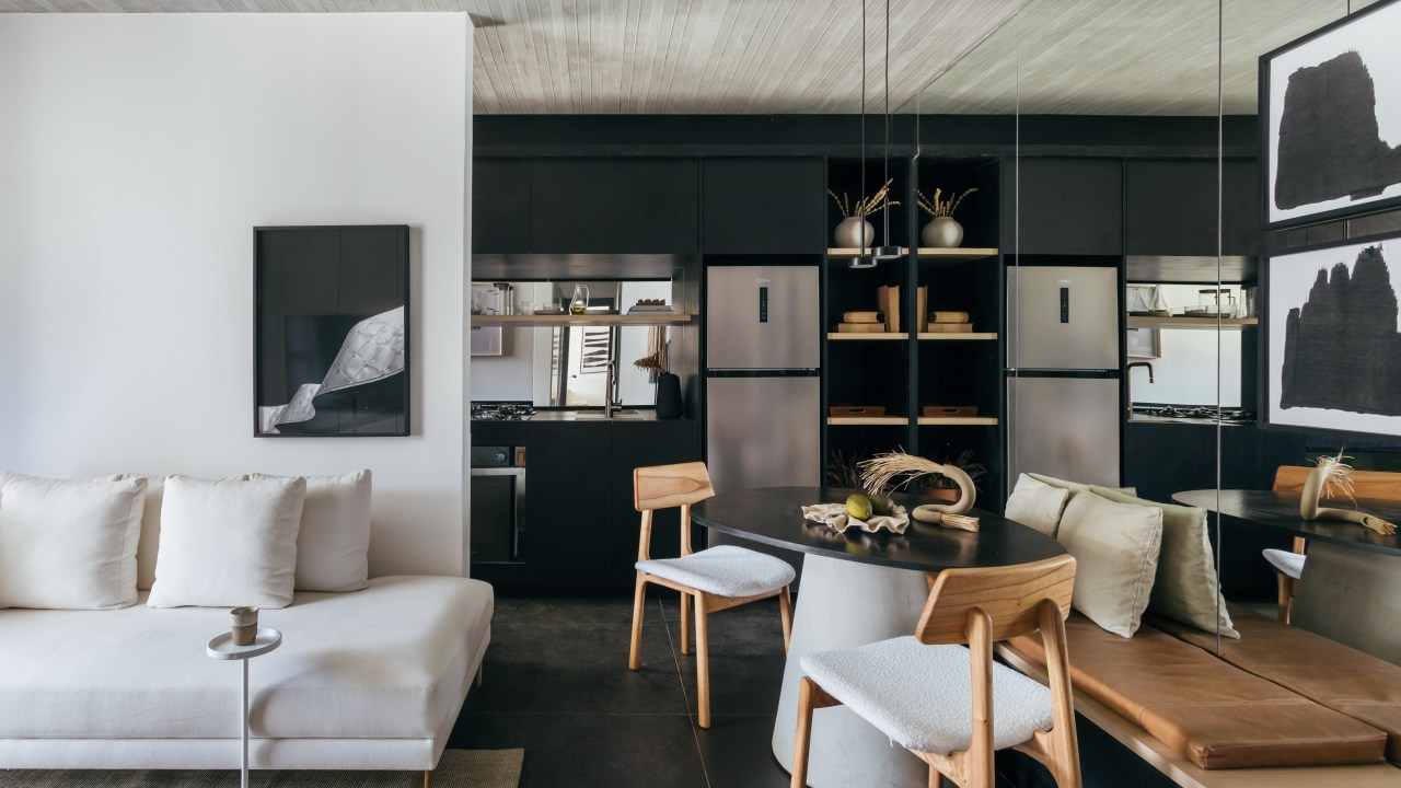 Apê de 73 m² tem marcenaria ebanizada e piso de porcelanato preto. Projeto de Studio Leandro Neves. Na foto, sala, parede preta, teto de concreto, sofá branco, mesa redonda, cozinha integrada.