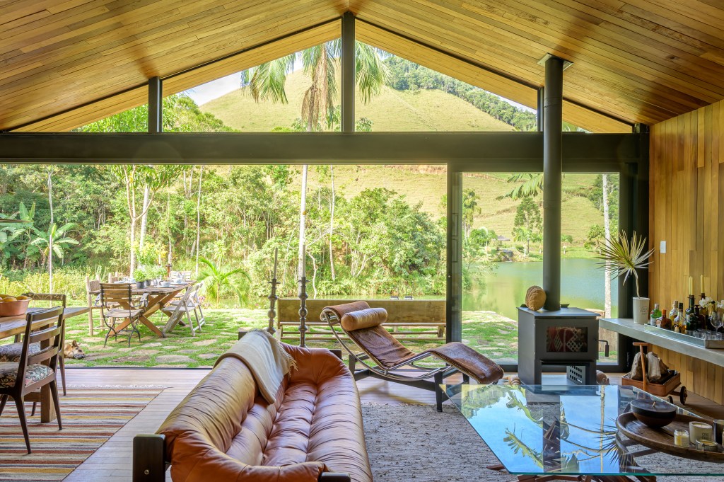 Depósito vira linda cabana no lago de 100 m², com duas suítes e varanda. Projeto de Roberto Souto Interiores. Na foto, sala, janela com vista para jardim, sofá de couro laranja.