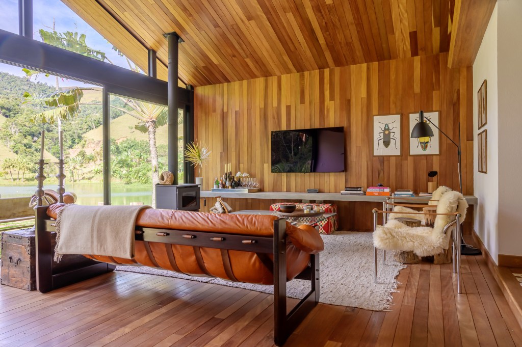 Depósito vira linda cabana no lago de 100 m², com duas suítes e varanda. Projeto de Roberto Souto Interiores. Na foto, sala, janela com vista para jardim, sofá de couro laranja.